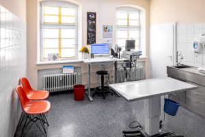 Ein Behandlungszimmer mit Computer, Untersuchungszubehör, einem Behandlungstisch in der Mitte und zwei Besucherstühlen.